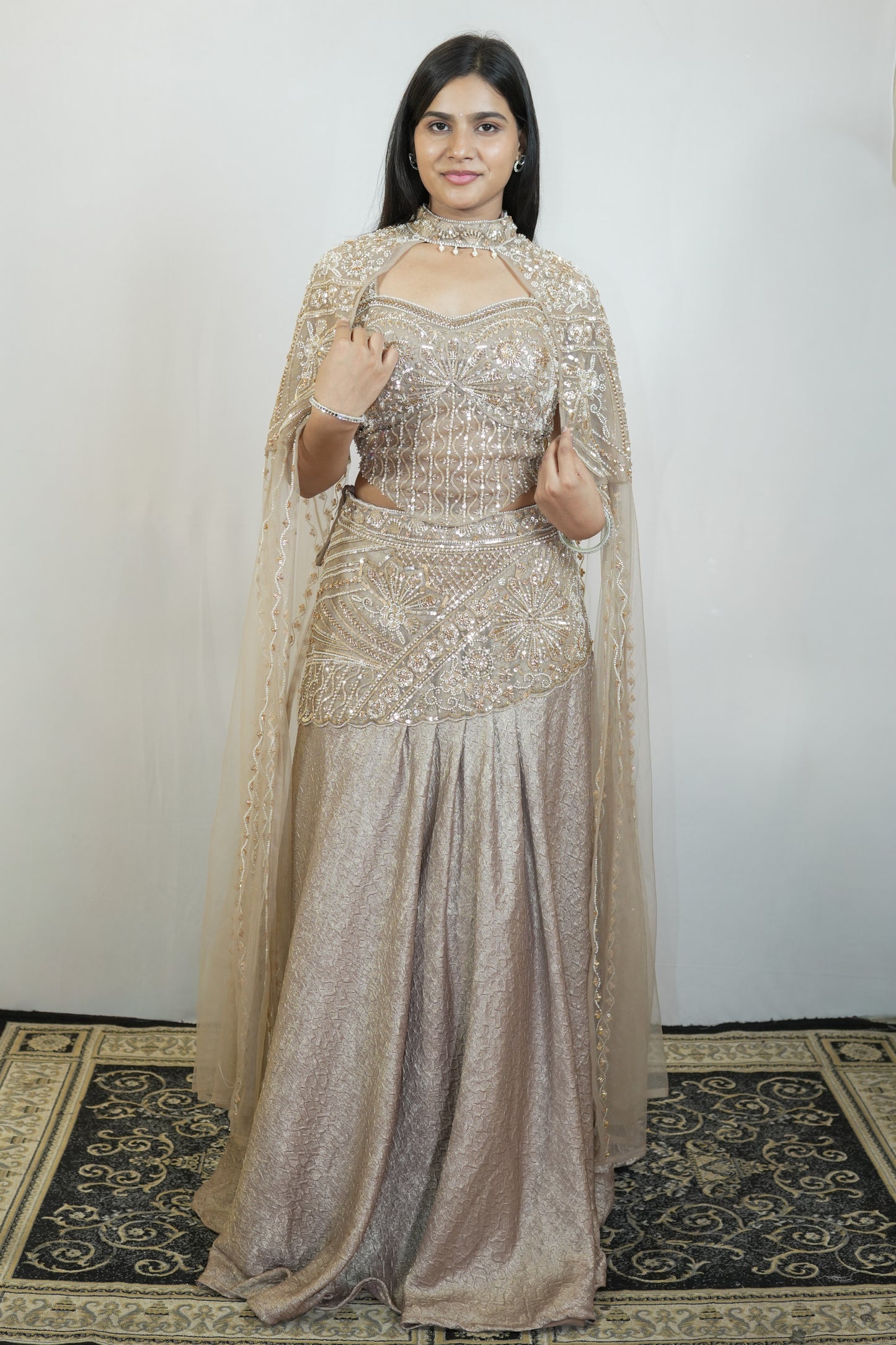A woman modeling a traditional Indian outfit consisting of a heavily embellished beige choli and a long pleated skirt with a matching sheer dupatta, set against a plain light gray wall and a patterned rug.