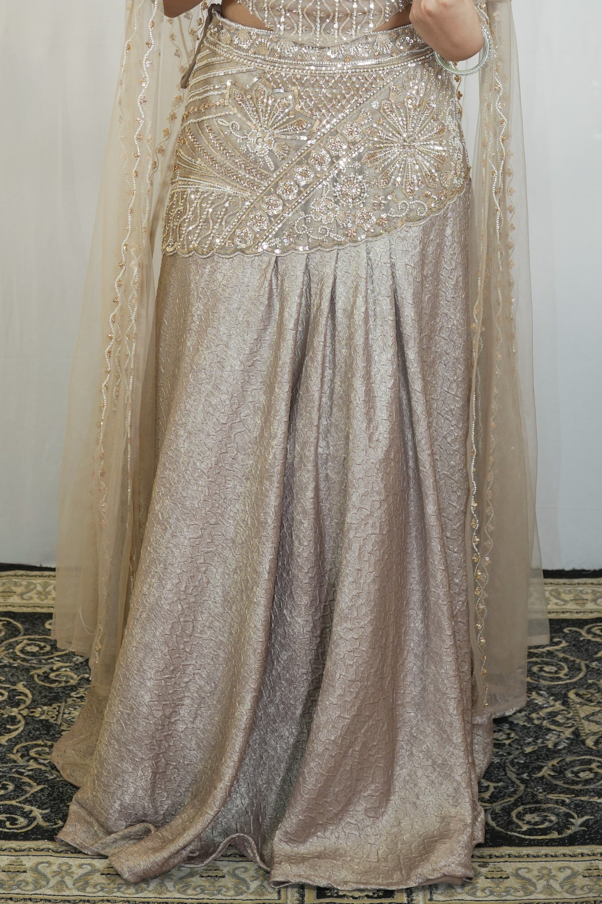 A close-up of a traditional Indian outfit featuring a heavily embellished beige and gold skirt with a matching sheer dupatta, worn by a model standing on a patterned rug.