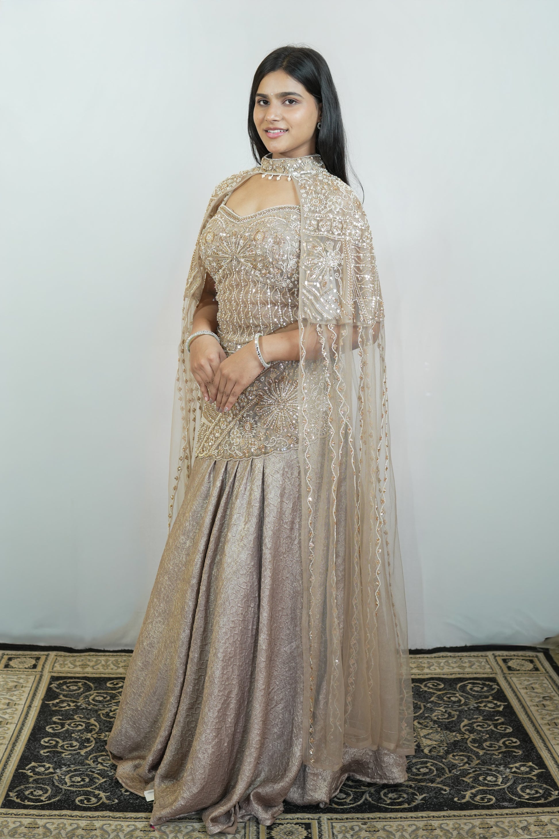 A woman modeling a traditional Indian lehenga choli in a champagne beige color with intricate gold embroidery, standing on a patterned rug against a plain light gray wall.