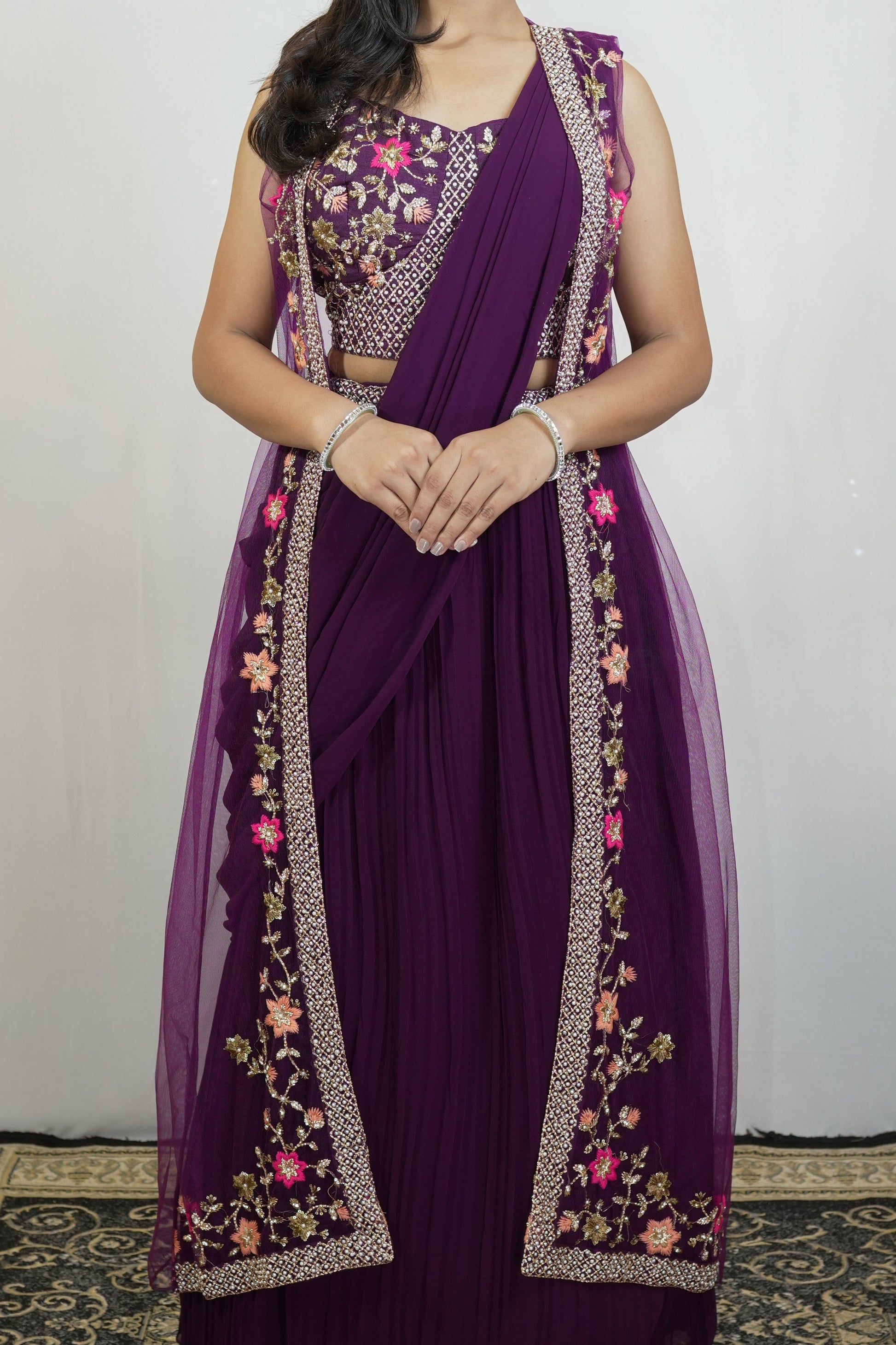 A woman wearing a deep purple traditional outfit with floral embroidery and metallic border details, standing against a plain light wall.