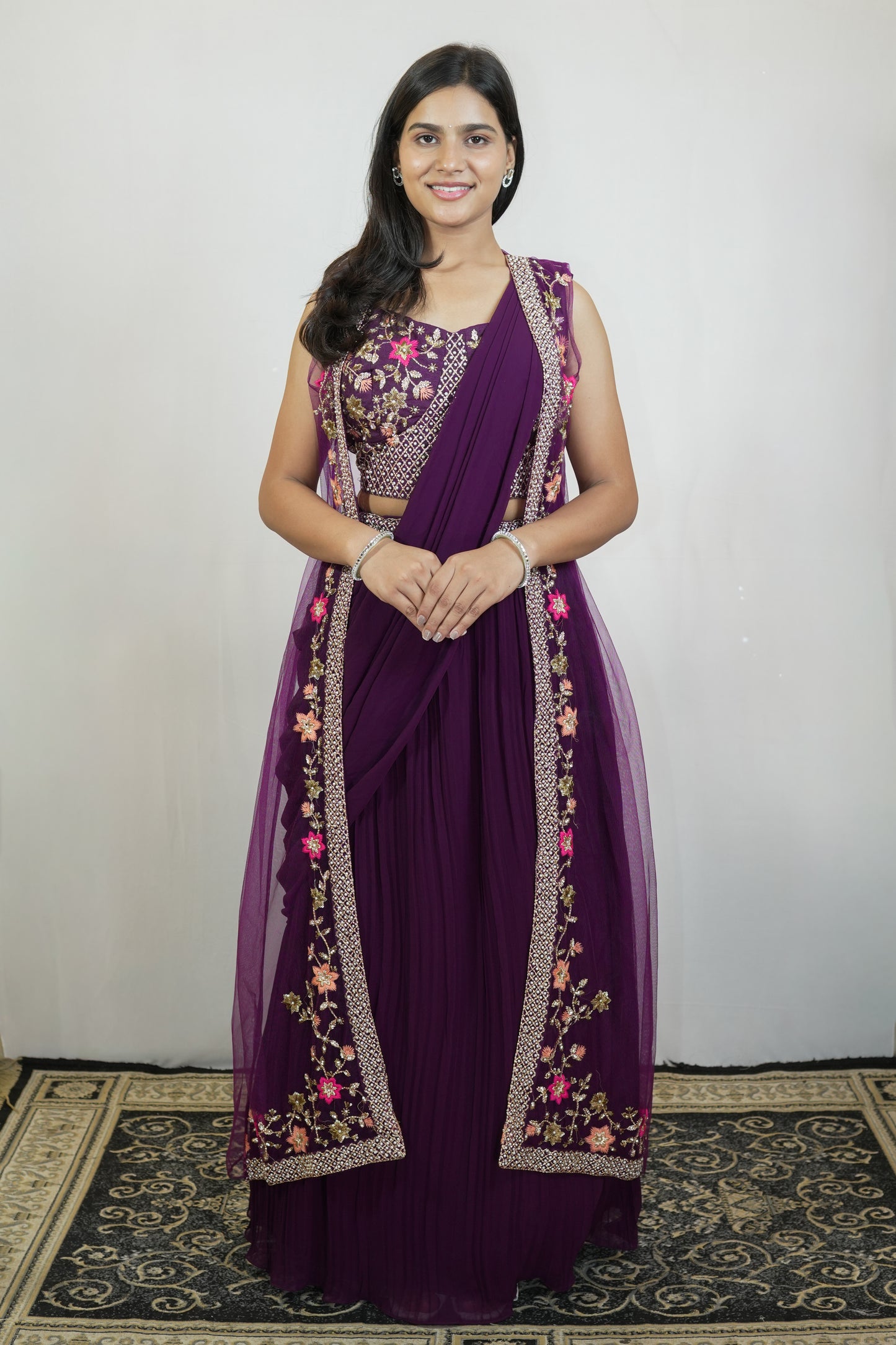 A woman modeling a traditional Indian outfit consisting of a deep purple georgette skirt, a matching embroidered sleeveless blouse, and a sheer purple dupatta draped over her shoulders, set against a plain light gray wall.