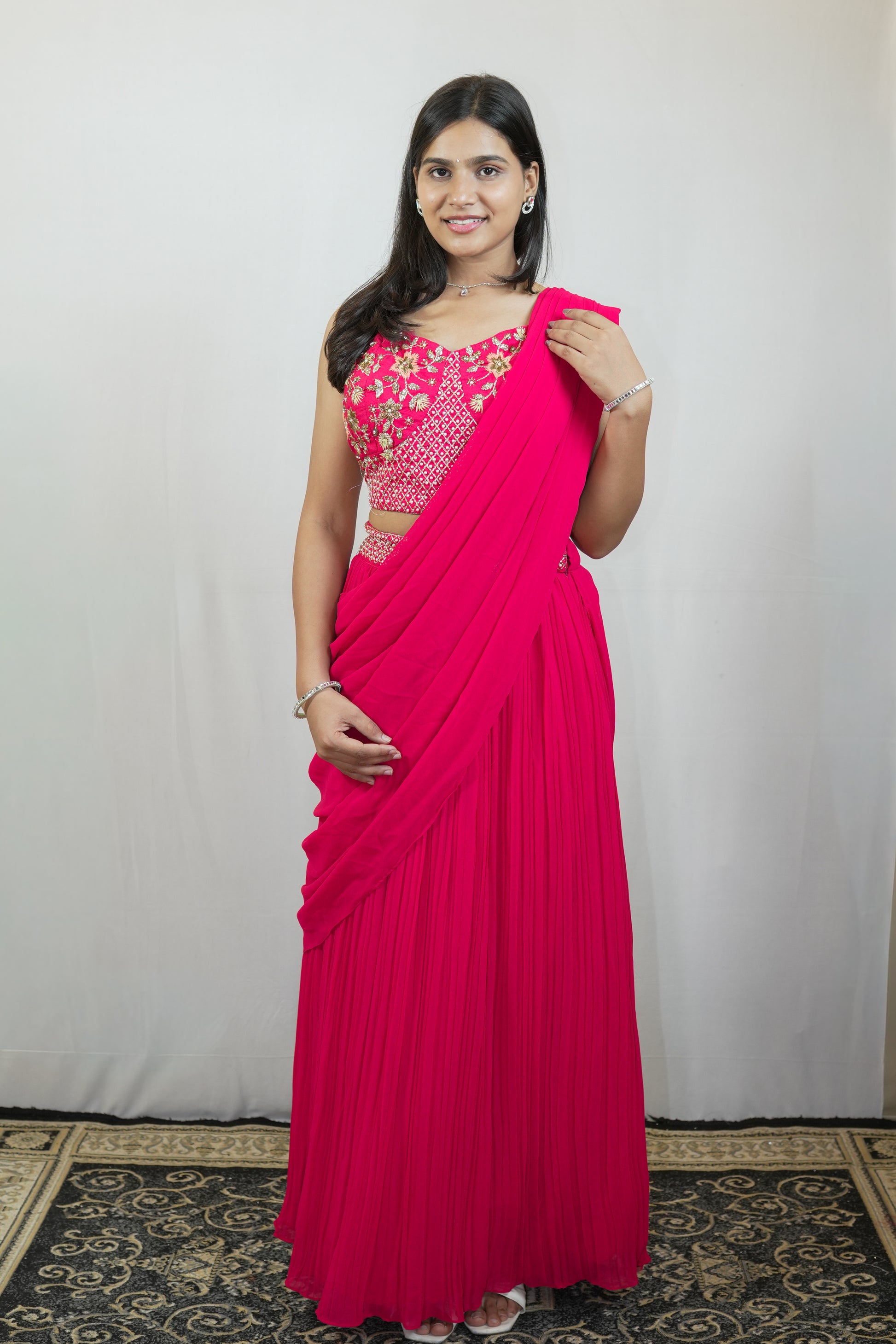 A woman modeling a bright pink pleated sari with a matching pink embroidered blouse, standing against a light gray fabric backdrop.