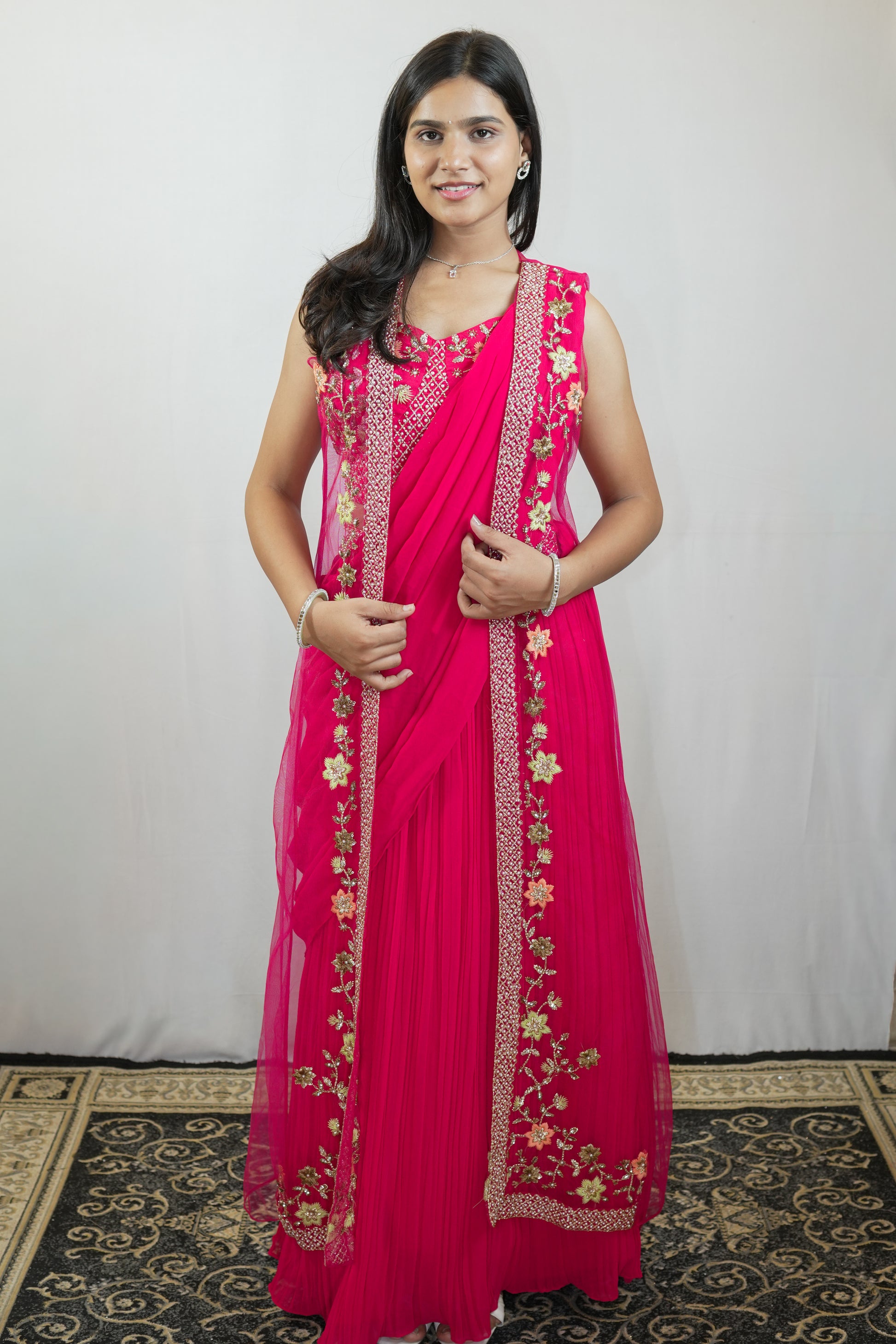 A woman modeling a bright pink traditional dress with gold floral embroidery and a matching sheer dupatta, standing against a plain light gray backdrop.