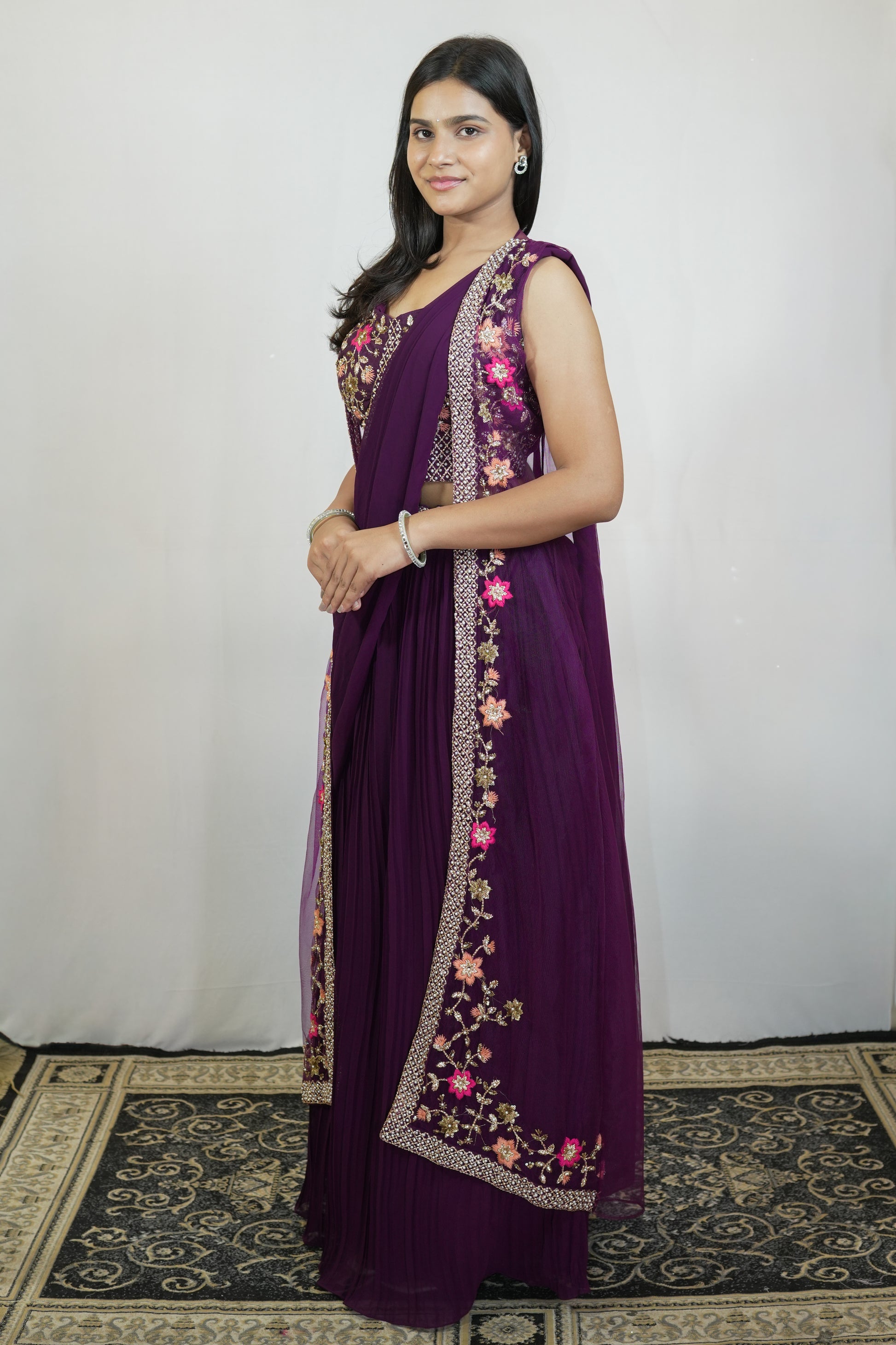 A woman modeling a deep purple traditional dress with floral embroidery and a matching embroidered dupatta, standing against a white backdrop.