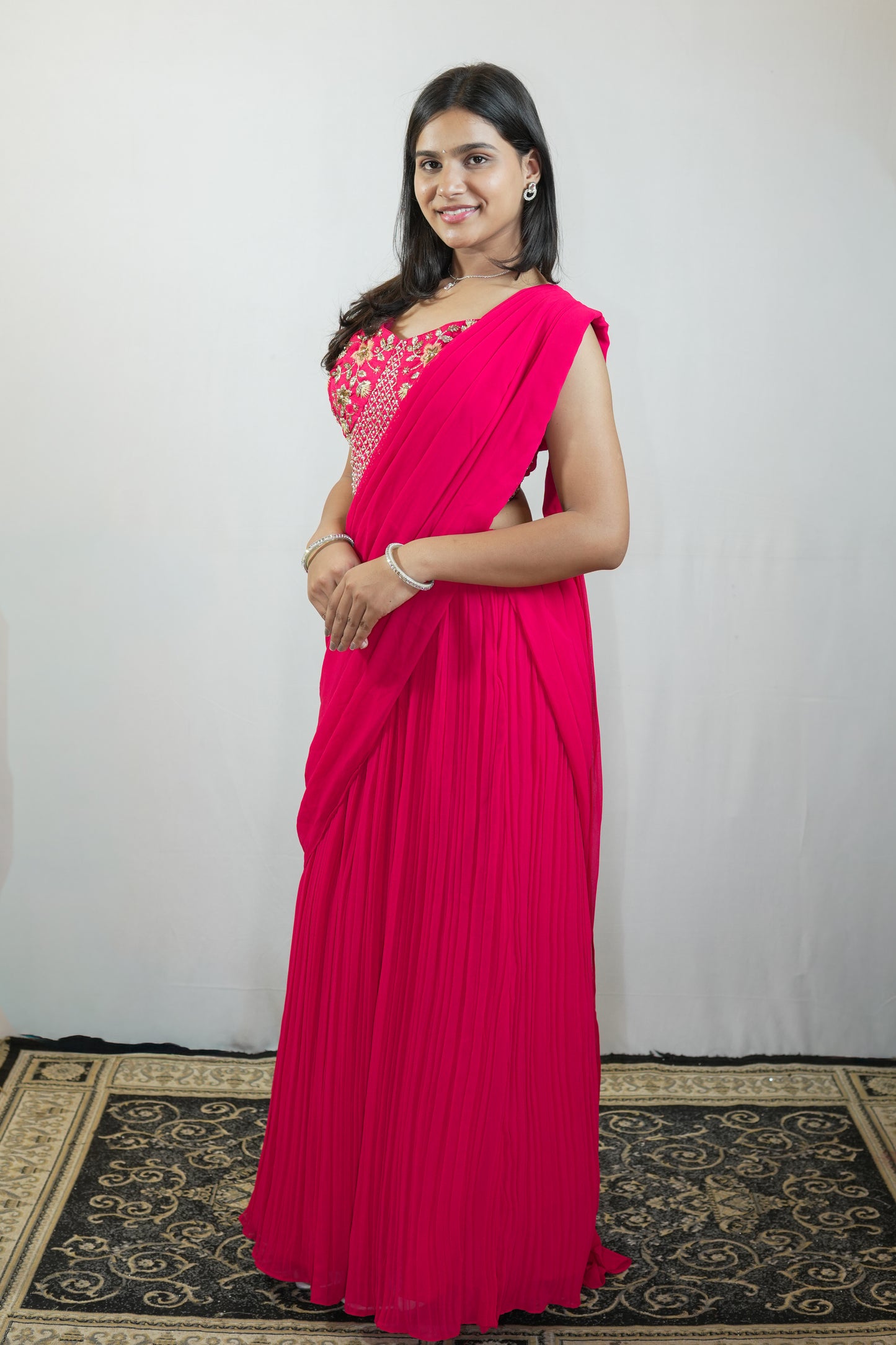 A woman wearing a bright pink pleated sari with a heavily embroidered pink and gold sleeveless blouse, standing on a patterned rug against a plain light gray backdrop.