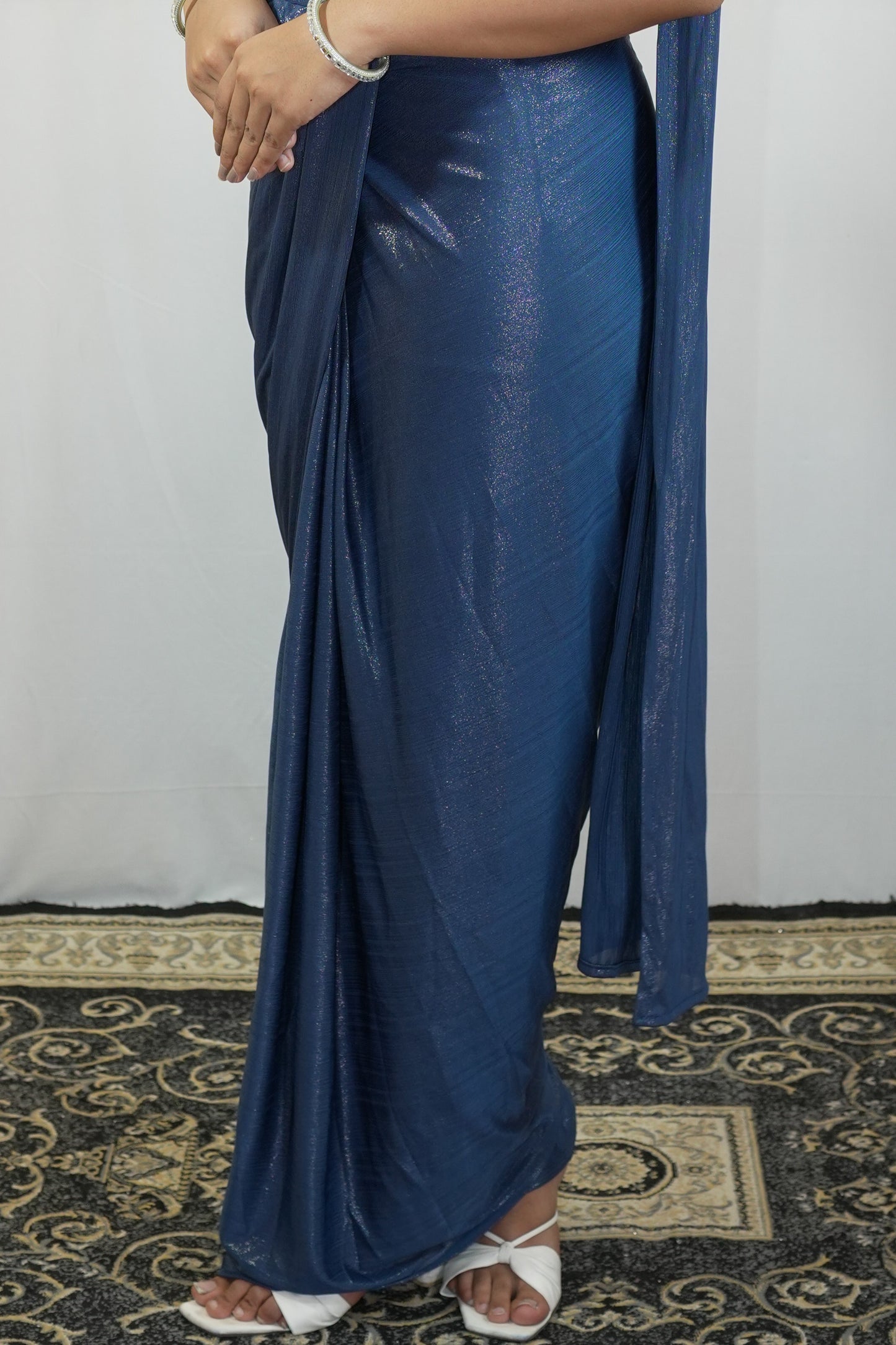 A person wearing a metallic blue saree with a matching dupatta, paired with white sandals, standing on a patterned rug against a white backdrop.
