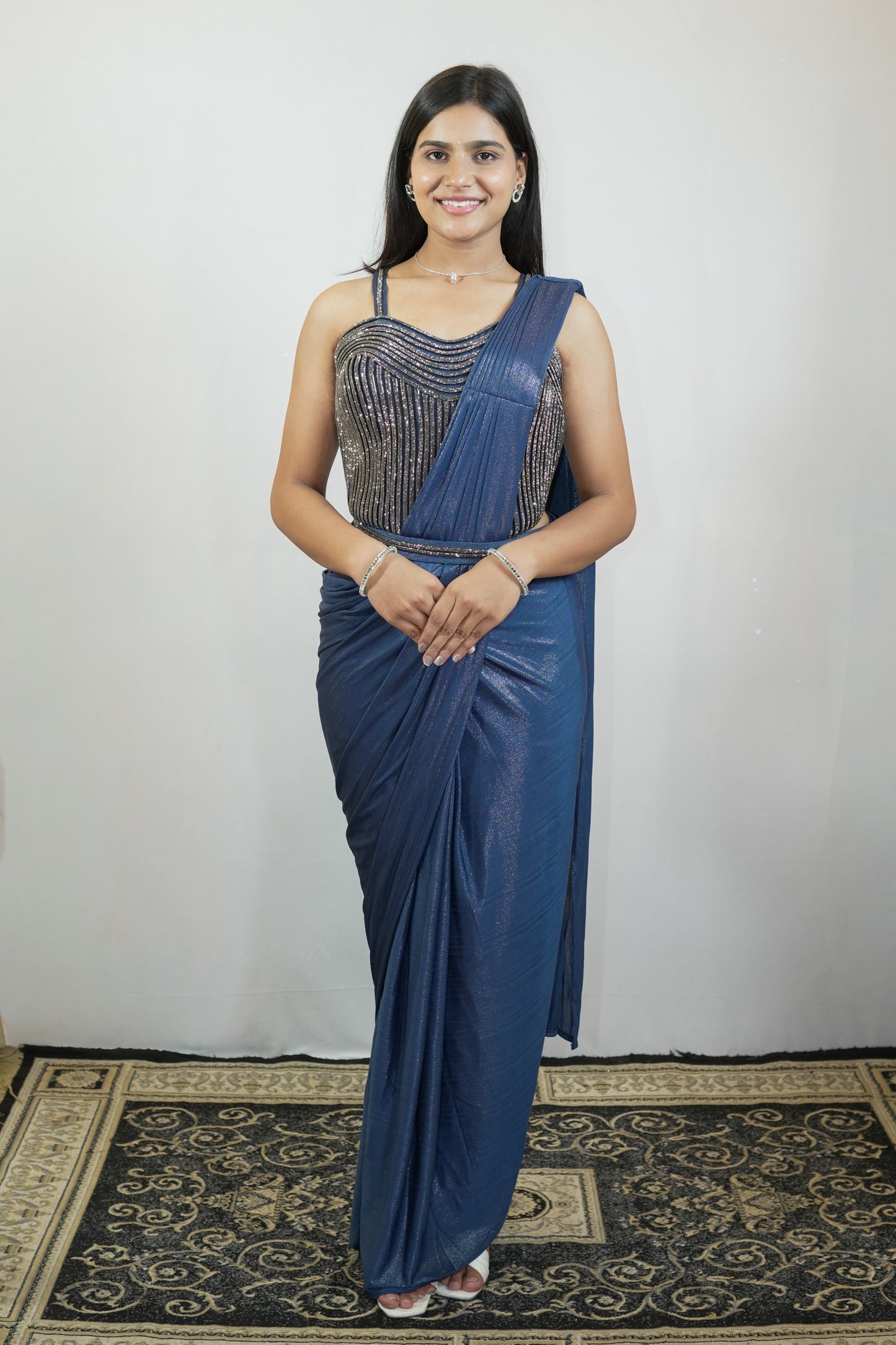 A woman modeling a blue saree with a heavily embellished silver sequined bodice, standing against a plain light gray wall on a patterned rug.