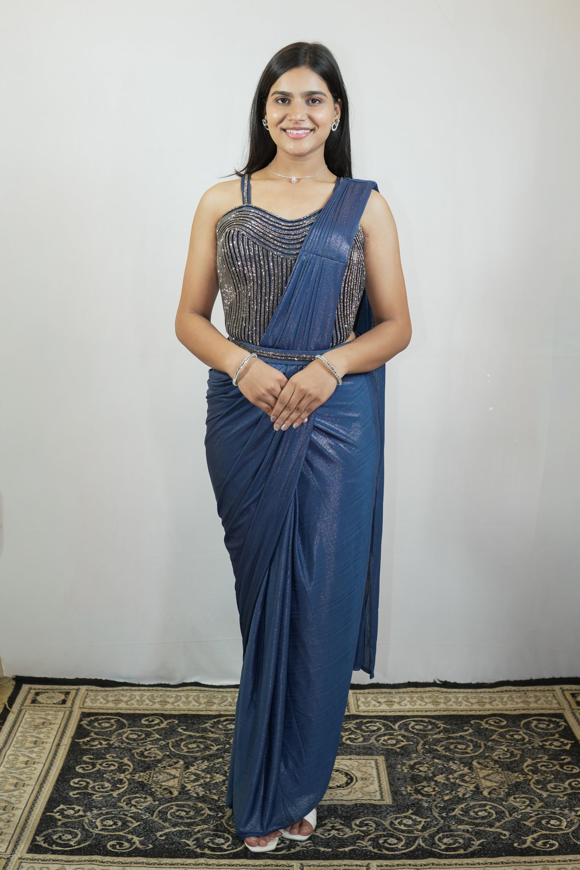 A woman modeling a blue saree with a heavily embellished silver sequined bodice, standing against a plain light gray wall on a patterned rug.